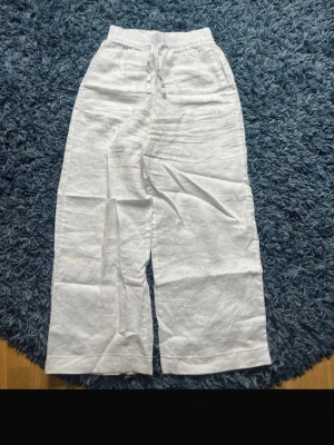 Vita vida linnebyxor med dragsko - Säljer ett par vita linnebyxor med bred passform och resår i midjan samt dragsko för skön och justerbar fit. Byxorna är raka i modellen och har en avslappnad look, perfekta för sommardagar. Materialet är luftigt linne som ger en fräsch känsla.