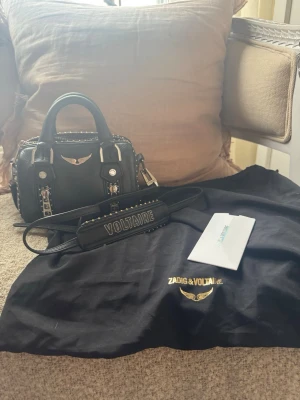 Svart handväska Zadig & Voltaire - Snygg svart handväska från Zadig & Voltaire med silverdetaljer, kedjor och nitar. Väskan har både handtag och avtagbar axelrem med logotypen 'VOLTAIRE'. Jag har kvar påsen och pris lappen från när jag köpte den. Den är i fint skick de ända är att den är lite rostig på ena dragkedjan men annars är den som ny🥰🥰
