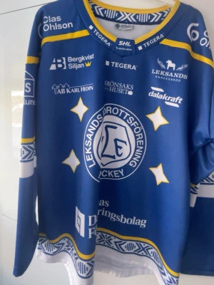 Leksands IF hockeytröja blå Bauer - Säljer en blå Leksands IF hockeytröja från Bauer med SHL-logga. Tröjan har vita och gula detaljer, tryckta sponsorer och klubbmärke på bröstet. Långärmad modell med rund hals och sportig design, perfekt för hockeyfans. Tröjan sitter som en s och är aldrig nästan Andvänd 