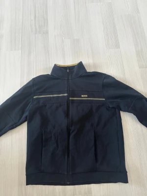 Marinblå jacka från BOSS - Snygg marinblå jacka från BOSS med hög krage och dragkedja framtill. Jackan har diskreta detaljer med tunna ränder över bröstet och BOSS-logga på vänster sida. Tillverkad i mjukt bomullsmaterial och har långa ärmar. Det är en limited edition så ganska ovanlig