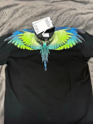 Marcelo Burlon svart t-shirt med vingar - Svart t-shirt från Marcelo Burlon med ikoniskt vingtryck i blått, grönt och gult runt halsen. Klassisk passform och rund hals. Perfekt statement-plagg för dig som vill sticka ut.
