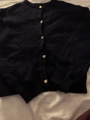 Blå kofta med hjärtknappar - Söt blå kofta med unika guldfärgade hjärtformade knappar framtill. Koftan har långa ärmar och ribbade muddar vid ärmslut och halsringning. Perfekt för dig som gillar detaljer och vill ha något extra i garderoben.