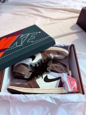 Nike Air Jordan 1 Travis Scott High - Säljer ett par Nike Air Jordan 1 Travis Scott High med bruna, vita och vinröda paneler, svart swoosh och detaljer. Skorna har högt skaft, svart snörning och kommer med extra skosnören. Materialet är en mix av mocka och läder.