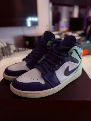 Air Jordan 1 Mid blå/grön/vit - Snygga Air Jordan 1 Mid sneakers i blått, vitt och mintgrönt. Klassisk siluett med perforerad tåbox, blå swoosh och detaljer i läder. Skön passform och ikonisk Jordan-logga på sidan. Perfekta för dig som vill sticka ut med stilrena färgblock.