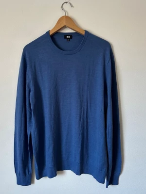 Blå stickad tröja från Uniqlo - En stilren blå stickad tröja från Uniqlo med rund halsringning och långa ärmar. Tröjan har ribbade muddar vid ärmslut och nederkant, vilket ger en clean och klassisk look. Perfekt att matcha med jeans eller chinos. 8D12