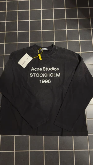 Svart långärmad topp Acne Studios - Svart långärmad topp från Acne Studios med vit text 'Acne Studios STOCKHOLM 1996' på bröstet. Klassisk rund halsringning och stilren design. Perfekt för dig som gillar minimalistiskt mode och vill ha en statement-piece från ett populärt svenskt märke.