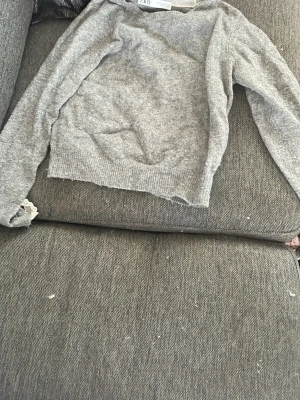 Grå stickad tröja från Zara - Mysig grå stickad tröja från Zara med rund halsringning och långa ärmar. Tröjan har ribbstickade muddar vid ärmslut och nederkant samt en söt spetsdetalj i halsen. Perfekt för lager på lager och enkel att matcha.