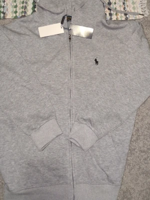 Grå zip-hoodie från Polo Ralph Lauren  - Snygh hoodie från Polo med dragkedja framtill och justerbar huva med snören. Klassisk modell med ribbade muddar vid ärmslut och nederkant. Perfekt för en chill och avslappnad stil. Riktigt fräsch och tjejerna kommer beundra dig i skolan kan jag lova. Sprillans ny.