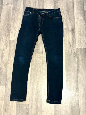 Nudie Jeans Skinny Lin - Snygga mörkblå jeans från Nudie Jeans som är som helst nya då de knappt är använda. Modellen är slim fit med raka ben och har detaljerade bakfickor med Nudie-signaturen. Perfekta för dig som gillar stilrena och tidlösa jeans. Dry Deep Orange heter denna modell. 30/30 i storlek. 