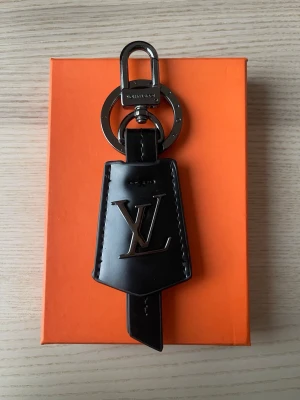 Vuitton svart nyckelring - Snygg svart nyckelring från  Vuitton i läder med metalliska detaljer och LV-logga framtill. Har en robust karbinhake och ring i silverfärgad metall. Perfekt att hänga på väskan eller använda till nycklar för en lyxig touch.