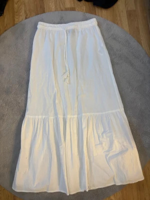 Vit långkjol med volang och snörning - Säljer en vit långkjol med volangkant och snörning i midjan. Köpt för 400kr. Kjolen har en avslappnad passform och är gjord i ett lätt bomullstyg, perfekt för varma dagar. Enkel och stilren design som passar till många olika looks. Använd en gång då den är för lång för mig (är 163cm) 