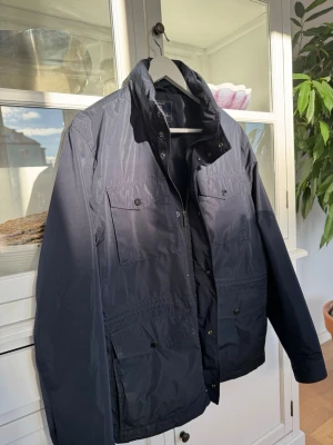 Mörkblå vindjacka med fickor - Snygg mörkblå vindjacka med flera praktiska fickor framtill och hög krage. Jackan har dragkedja och knappar, samt justerbar midja för bättre passform. Perfekt för kyliga dagar och enkel att matcha med olika outfits. Väldigt lite använd och väldigt bra skick! NYPRIS 1900.