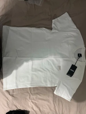 Vit t-shirt från BOSS med små guld detaljer - Klassisk vit t-shirt från BOSS  med rund hals och diskret BOSS-logga på bröstet. T-shirten har korta ärmar och är gjord i mjuk bomull för en skön och stilren look. Perfekt till jeans eller shorts för en clean outfit.