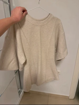 Beige oversized topp med vida ärmar - Trendig beige topp med oversized passform och vida, halvlånga ärmar. Tillverkad i ett mjukt, tjockare material med dekorativa sömmar framtill. Rund halsringning och minimalistisk design som passar till många stilar.