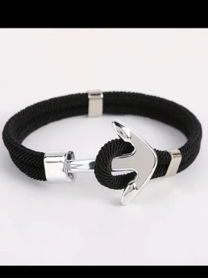 Svart armband med ankardetalj - Stilrent svart armband med en blank silvrig ankare som lås. Armbandet är gjort av flätat repmaterial och har metalliska detaljer som ger en modern och nautisk känsla. Perfekt accessoar för dig som gillar marina vibbar och enkel design.