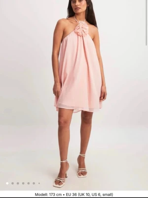 Ljusrosa chiffongklänning med blomdetalj - Supersöt ljusrosa kortklänning i chiffong med en stor blomdetalj framtill vid halsen. Klänningen är ärmlös och har en luftig, A-linjeformad passform som ger en romantisk vibe. Perfekt för dig som vill sticka ut med en feminin look. Ny med prislapp kvar☺️