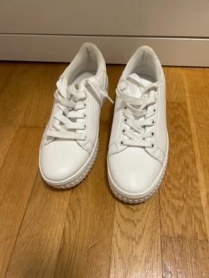 Vita sneakers med gul detalj - Snygga vita sneakers med tjock sula och en tunn gul linje på sidan. Skorna har klassisk snörning och är tillverkade i ett slätt syntetmaterial. Perfekta för dig som gillar en clean look med en liten färgklick.