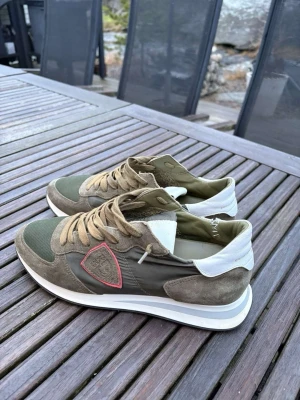 Olivgröna sneakers från Philippe Model - Snygga sneakers i olivgrönt och beige från Philippe Model med mockadetaljer, vit sula och vitt parti vid hälen. Skorna har en ikonisk sköldlogga på sidan med röd kant och klassisk snörning. Perfekta för en trendig streetwear-look.