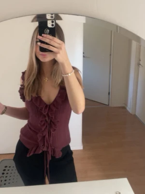 Vinröd blus med volang och snörning - Supersnygg vinröd blus med volangdetaljer och snörning framtill från Zara. Toppen är ärmlös och har en figurnära passform. Använd ca 2 gånger. Mycket bra skick. Säljs inte längre på Zaras hemsida.