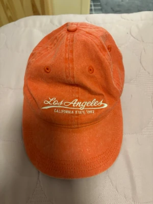 Orange keps Los Angeles brodyr - Snygg orange keps med vit brodyr där det står 'Los Angeles California State 1992' på framsidan. Klassisk böjd skärm och justerbar passform bak. Tillverkad i bomullstyg, perfekt för en avslappnad streetwear-look.