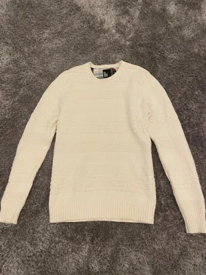 Vit stickad tröja från Red Collar Project - Klassisk vit stickad tröja från Red Collar Project med rund halsringning och diskret strukturmönster. Tröjan har långa ärmar och ribbade muddar vid ärmslut och nederkant. Perfekt för lager på lager och enkel att matcha med olika stilar.