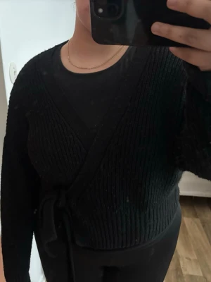 Svart omlottstickad tröja - Säljer en svart stickad omlott-tröja med knytband i midjan. Tröjan har ribbstickad struktur, långa ärmar och en djup v-ringning. Perfekt för lager på lager och enkel att styla till olika outfits.