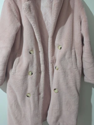 Ljusrosa teddyjacka med knappar - Supersöt ljusrosa teddyjacka med dubbelknäppning i guldiga knappar och två fickor på sidorna. Jackan har en mjuk, fluffig yta och quiltat foder på insidan. Perfekt för dig som vill ha en mysig och trendig look i höst och vinter.