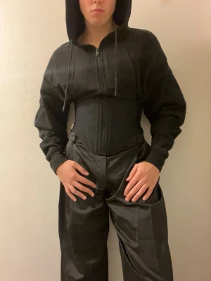 H&M x Mugler huvtröja - Säljer en svart H&M x Mugler-hoodie med dragkedja framtill och huva med snören. Hoodien har en korsettmidja och långa ärmar med resår. Materialet känns mjukt och bekvämt, troligen bomullsblandning.