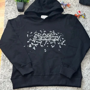 Svart hoodie Maison Margiela - Svart hoodie från Maison Margiela med ikoniskt siffror-tryck och gråa färgstänk på bröstet. Klassisk huva, ribbade muddar och avslappnad passform. Perfekt för dig som gillar streetwear och vill sticka ut med en unik design.