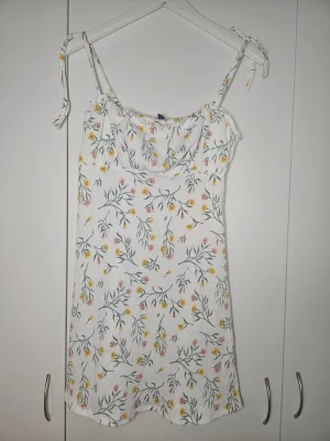 Vit blommig sommarklänning H&M Divided - Söt vit kortklänning från H&M Divided med blommigt mönster i gult och rosa. Klänningen har smala axelband med knyt och en lätt, luftig känsla. Perfekt för varma dagar och enkel att styla med sneakers eller sandaler.