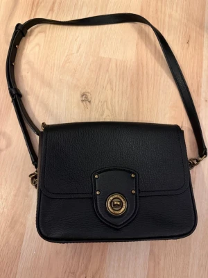 Svart axelväska i läder från Ralph Lauren - Snygg svart axelväska från Ralph Lauren i äkta läder med justerbar rem och guldfärgade metalldetaljer. Klassisk rektangulär form och lock med vridlås framtill. Perfekt storlek för det viktigaste och stilren design som passar till allt. Storlek 20x15.