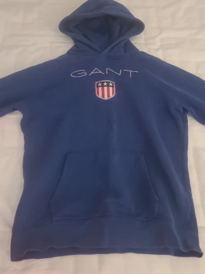 Blå hoodie från GANT med logga - Blå hoodie från GANT med vit broderad logga och ett sköldmärke med stjärnor och ränder på bröstet. Klassisk känguruficka framtill och stor huva. Tillverkad i mjukt bomullsmaterial, perfekt för en avslappnad och sportig stil.