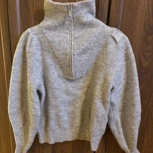 Grå stickad tröja med puffärm - Mysig grå stickad tröja med hög krage och dragkedja. Tröjan har trendiga puffärmar och ribbstickade detaljer vid ärmslut och nederkant. Perfekt för dig som gillar en stilren men ändå unik look.