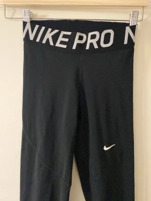 Svarta Nike Pro leggings - Snygga svarta Nike Pro leggings med bred resår i midjan där det står 'NIKE PRO' i vitt. Stretchigt och mjukt material som sitter tight och bekvämt. Perfekta för träning eller chill. Klassisk vit Nike-logga på benet.