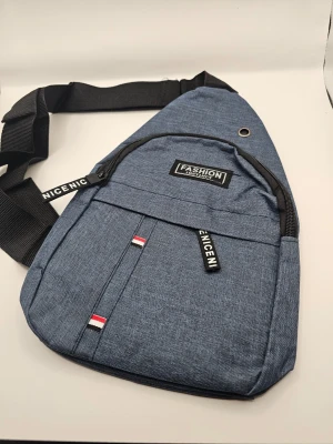 Blå crossbody-väska Fashion Axelremsväska  - Snygg blå crossbody-väska med svart justerbar axelrem och dragkedja. Väskan har detaljer med texten 'NICENICENICE' och små rödvita band. Praktisk ficka framtill och metallögla upptill. Tillverkad i slitstarkt tygmaterial.