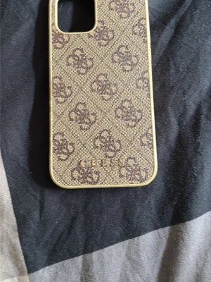 Beige mobilskal Guess monogram iPhone 12 Pro  - Snyggt beige mobilskal från Guess med klassiskt monogrammönster i brunt och guldfärgad logga längst ner. Skalet har en rektangulär form och är gjort i ett tåligt material med en lyxig känsla. Perfekt för dig som vill ha ett trendigt och stilrent skydd till mobilen.