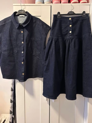 Mörkblå jeanssett, kjol och skjorta  - Säljer en mörkblå jeanssett, kjol och skjorta med korta ärmar och stora bröstfickor. Skjortan har klassisk krage och silverfärgade knappar framtill. Perfekt för en avslappnad och trendig look.