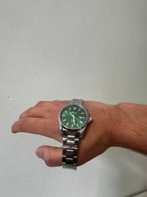 Grön klocka + plus Klockarmbandsverktyg - Snygg klocka  grön urtavla och datumvisning. Klockan har en klassisk rund form och en länk i silverfärgat stål som ger en clean och modern look. Perfekt accessoar för dig som vill sticka ut lite extra.