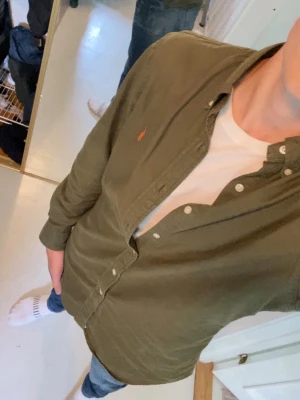 Skjorta från Ralph Lauren - Säljer en olivgrön skjorta från Ralph Lauren med klassisk knappslå och den ikoniska broderade loggan i röd på bröstet. Skjortan har lång ärm, normal passform och är tillverkad i mjuk bomull. Perfekt till jeans eller chinos för en clean look. Köpt för 2800 och är som ny, använt den 2 gånger och de syns knappt och därför tänkt sälja för 1850 men priset sitter inte i sten
