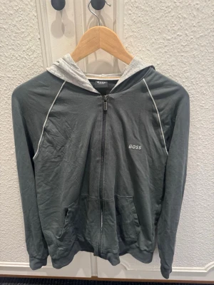 Grön zip hoodie från BOSS - Mörkgrå hoodie med dragkedja från BOSS. Tröjan har vit logga på bröstet, kontrasterande ljusgrå huva och vita sömdetaljer längs ärmarna. Tillverkad i mjukt material och har fickor framtill. Perfekt för en avslappnad och stilren look. Nypris 899