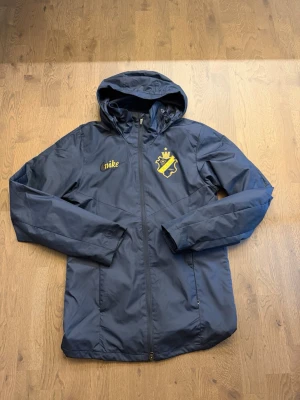 AIK Royal Edition Regnjacka  - Snygg mörkblå vindjacka från Nike med AIK-logga och huva. Jackan har dragkedja framtill och två sidofickor. Perfekt för dig som vill visa ditt AIK-stöd och hålla dig skyddad mot vinden. Gult Nike-tryck på bröstet och stor AIK-emblem.
