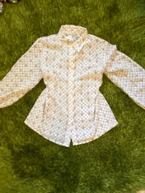 Vit mönstrad blus med krage - Snygg vit blus med rutigt mönster och små röda detaljer. Klassisk krage och knäppning framtill. Figursydd modell som ger en fin siluett och lätt tyg som också ger solskydd. 