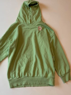 Grön hoodie med blompatch - Snygg ljusgrön hoodie med huva och ribbade muddar. På bröstet sitter en liten broderad blompatch i rosa och vitt som ger en söt detalj. Hoodien är i mjuk bomull och har en avslappnad passform, perfekt för chill dagar.