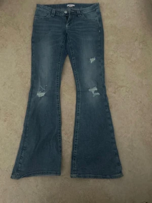 Blå bootcut jeans med slitningar - Säljer ett par blå bootcut jeans med slitna detaljer på framsidan och broderade fickor bak. Jeansen har klassisk femficksdesign och är tillverkade i denim. Perfekta för dig som gillar en retro och avslappnad stil.