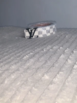 Vit och grå bälte  - Säljer ett snyggt bälte  i vitt och grått med klassiskt rutigt mönster och  i metall framtill. Bältet är gjort i skinn och har en stilren design som sticker ut.