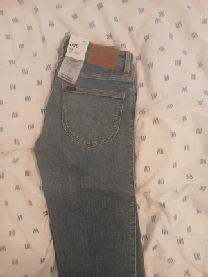 Blå raka jeans från Lee - Säljer ett par klassiska blå jeans från Lee i modellen 'Feels Like Indigo'. Jeansen har raka ben, fem fickor och snygga kontrastsömmar. Tillverkade i slitstark denim med Lee-logga på bakfickan och läderpatch i midjan. Perfekta för en avslappnad och trendig look. Den är helt ny från jens bolaget 