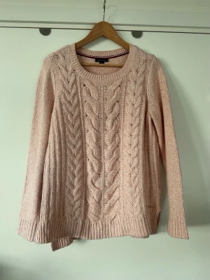Tommy Hilfiger - Ljusrosa kabelstickad tröja - storlek S - Klassisk stickad tröja från Tommy Hilfiger i storlek S i en ljusrosa melerad nyans. Tidlös modell med rund halsringning och långa ärmar. Framtill pryds den av ett flätstickat mönster som ger textur och detalj. Nederkanten och ärmsluten är ribbstickade. En liten metallbricka med loggan är fäst vid nederkanten. Fint skick!