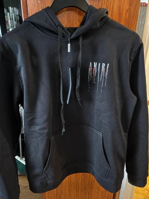Svart hoodie från Amiri med tryck - Svart hoodie från Amiri med stor huva och dragsko. Framtill finns ett mindre Amiri-tryck med färgstänk och på ryggen ett stort Amiri-tryck med droppande färgeffekt i vitt, blått och orange. Klassisk känguruficka och mjukt material, perfekt för en trendig streetwear-look.