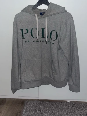 Grå hoodie från Polo Ralph Lauren - Snygg grå hoodie från Polo Ralph Lauren med stor grön logga på bröstet och vita dragsnören i huvan. Hoodien har känguruficka framtill och ribbade muddar vid ärmar och nederkant. Perfekt för en chill och stilren look.