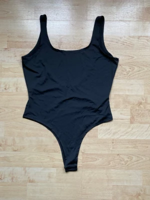 Svart basic body med breda axelband - En enkel och stilren svart body med breda axelband och rundad urringning både fram och bak. Tillverkad i ett stretchigt och mjukt material som sitter tajt mot kroppen. Perfekt att styla till olika outfits för en clean look.